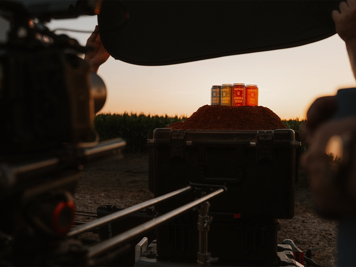 Cans of Rancho cocktails displayed on a mound of dirt during a sunset film shoot, with camera equipment in the foreground.