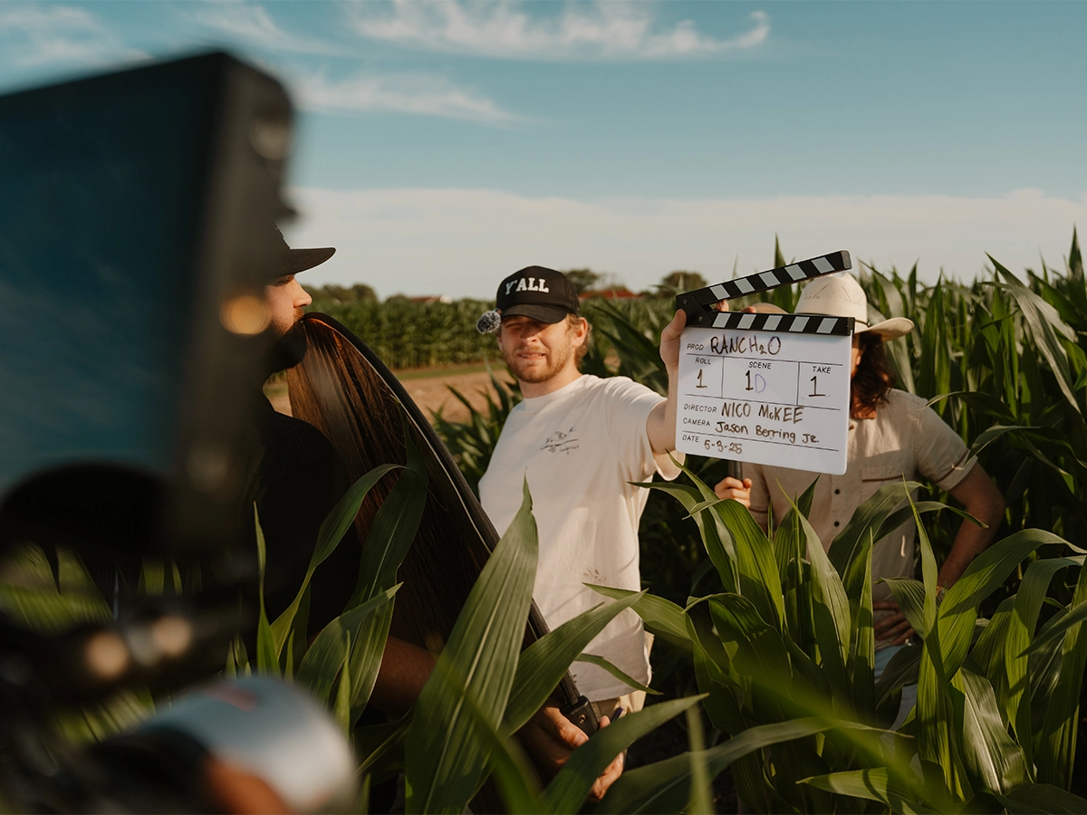 Film crew in a cornfield holding a clapperboard labeled Rancho H2O during an outdoor video shoot.