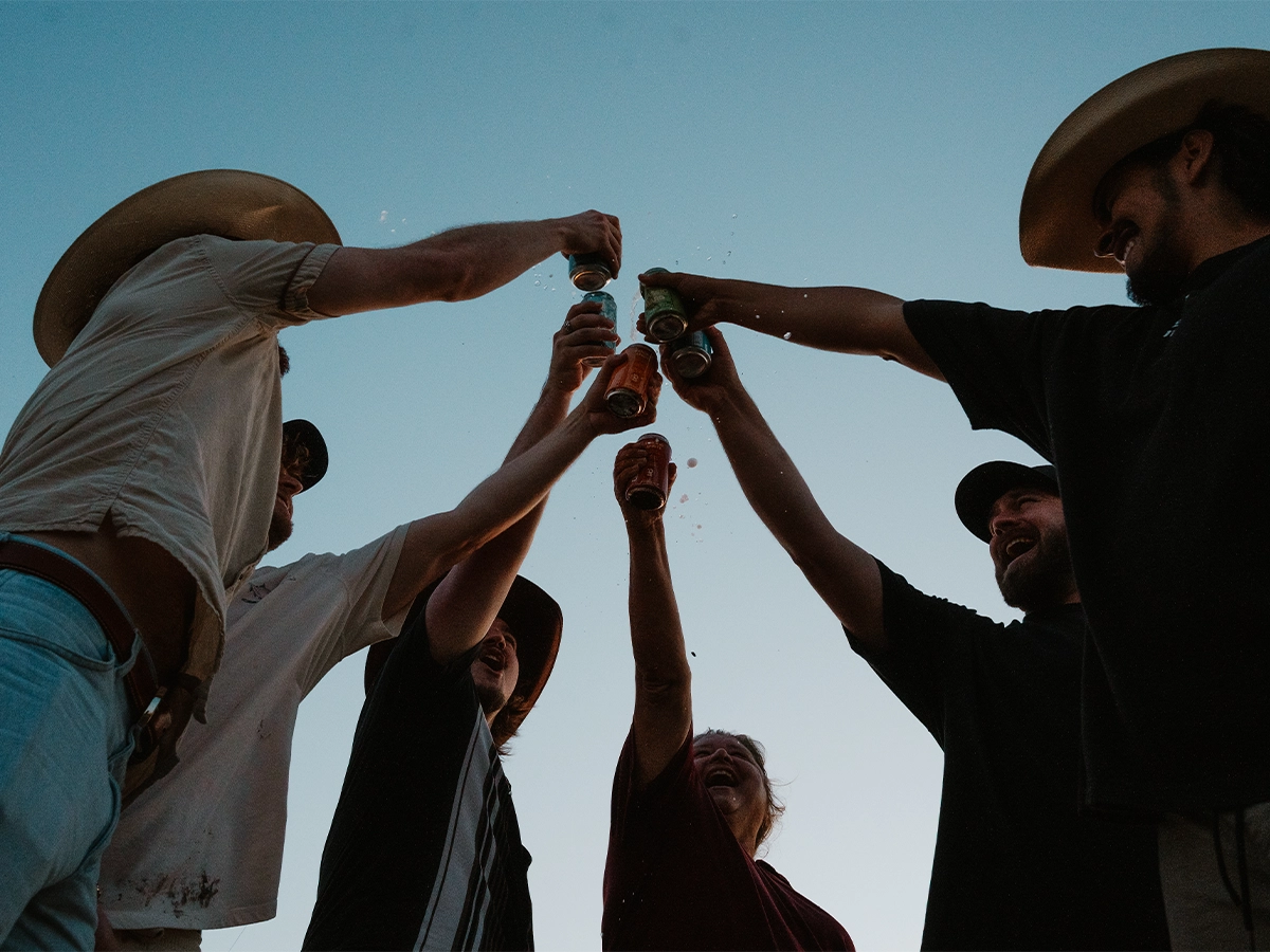 Group of friends in cowboy hats raising cans of Rancho cocktails in a celebratory toast against a clear sky.