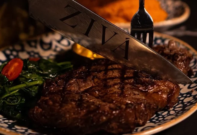 Close-up of a grilled steak served with sautéed vegetables, with a knife engraved ZIVÁ resting on top.